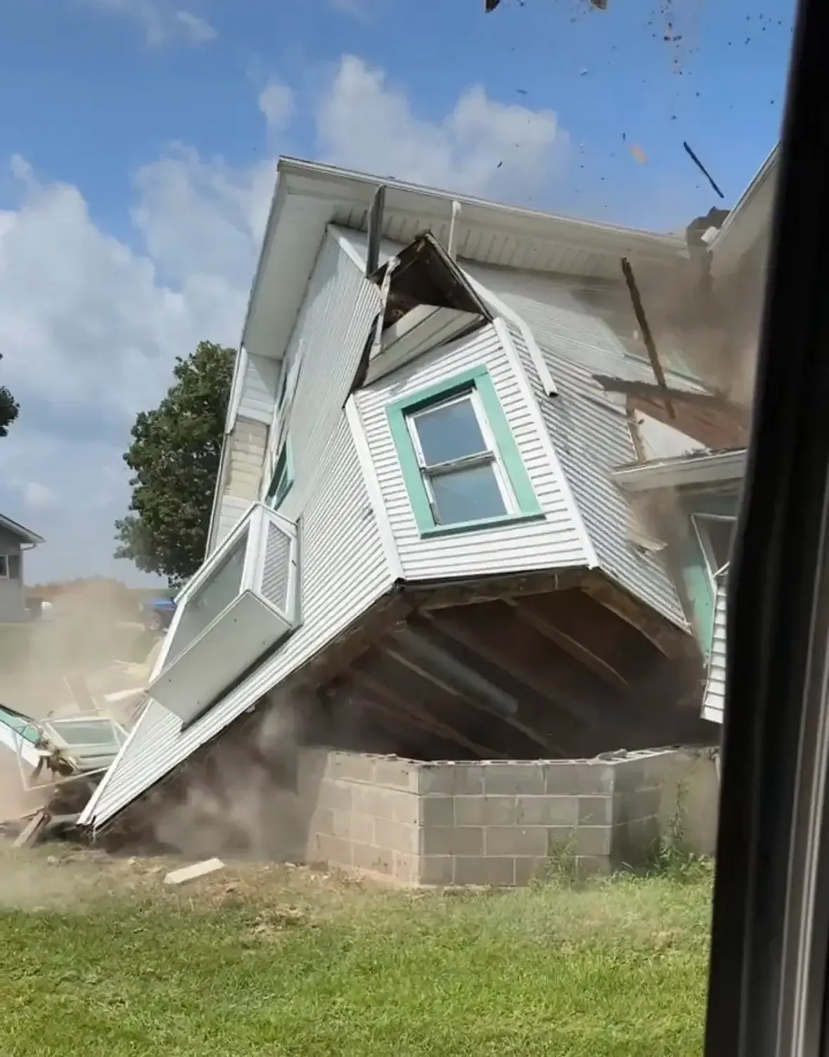 A two-story house tipping over during a Salz demolition, seen from the excavator cab