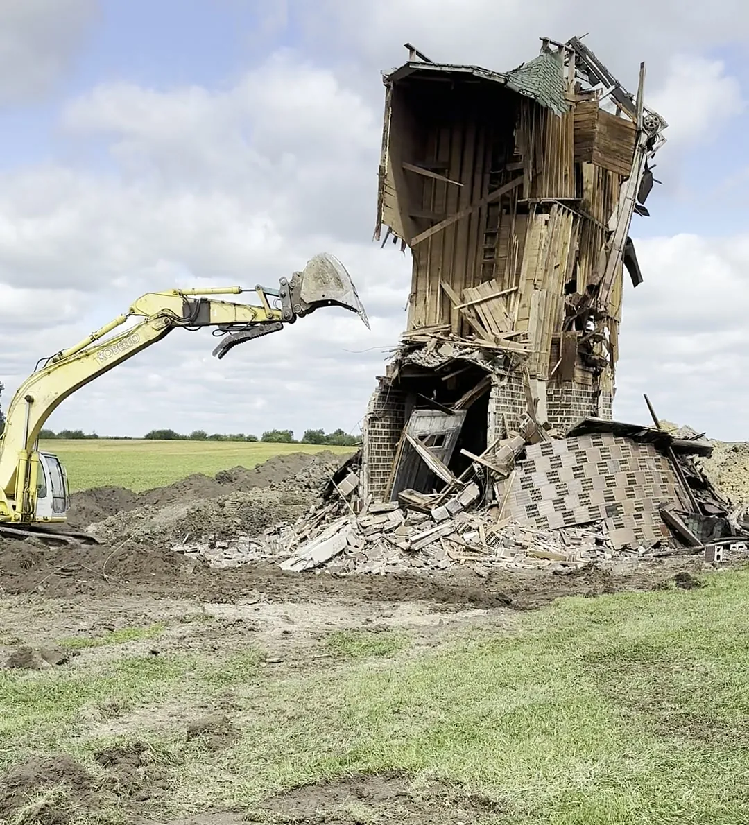 Yellow excavator pulling down the last wall of a brick farmhouse