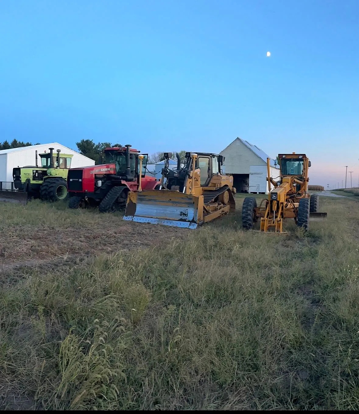 Salz tractors, dozer and grader lined up in a field at dusk beside white farm buildings
